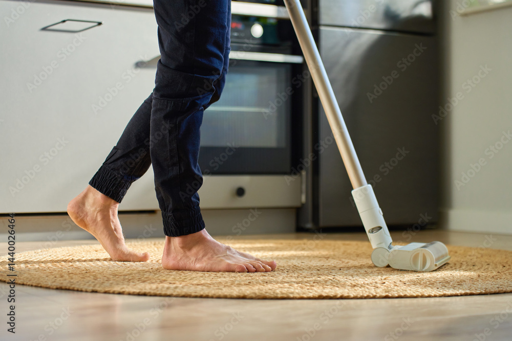 Barefoot man vacuuming round woven rug on wooden floor in kitchen. Person uses handheld stick vacuum cleaner in apartment. Concept of daily home cleaning and household chores