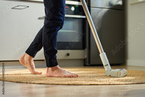 Obraz na płótnie Barefoot man vacuuming round woven rug on wooden floor in kitchen. Person uses handheld stick vacuum cleaner in apartment. Concept of daily home cleaning and household chores w salonie