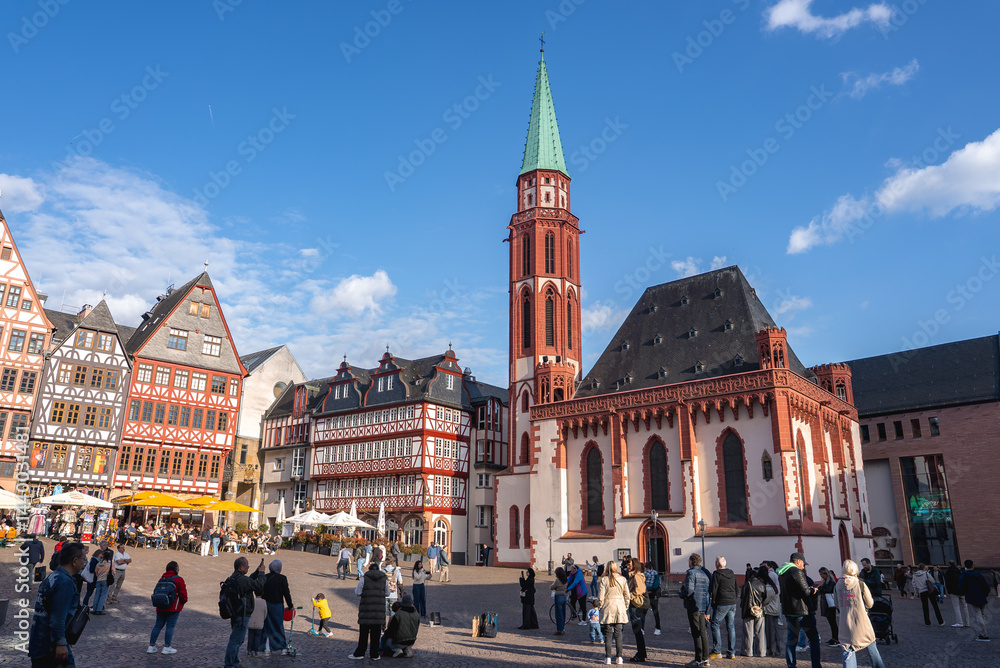 Romerberg square in Frankfurt, Germany, features Old St. Nicholas Church, half timbered buildings, and people enjoying the lively atmosphere under a blue sky.