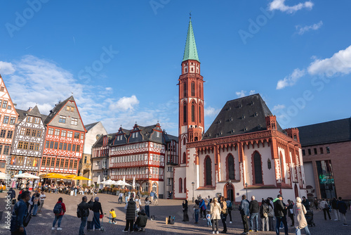 Obraz na płótnie Romerberg square in Frankfurt, Germany, features Old St. Nicholas Church, half timbered buildings, and people enjoying the lively atmosphere under a blue sky. w salonie