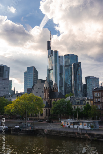 Obraz na płótnie Frankfurt's skyline features modern skyscrapers like Commerzbank Tower, a historic church with spires, lush trees, and the Main River with a riverside walkway. w salonie