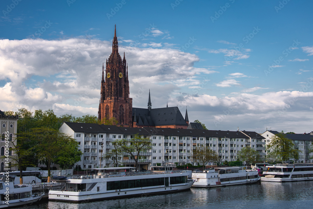 The Frankfurt Cathedral's Gothic spire rises against a partly cloudy sky, with the Main River, riverboats, residential buildings, and greenery in view.