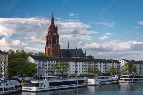 Obraz na płótnie The Frankfurt Cathedral's Gothic spire rises against a partly cloudy sky, with the Main River, riverboats, residential buildings, and greenery in view. w salonie