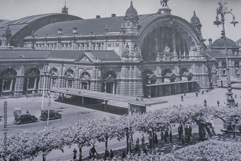 Frankfurt Central Station's neo Renaissance facade with a clock, decorative towers, tree lined street, vintage cars, pedestrians, and ornate street lamps.