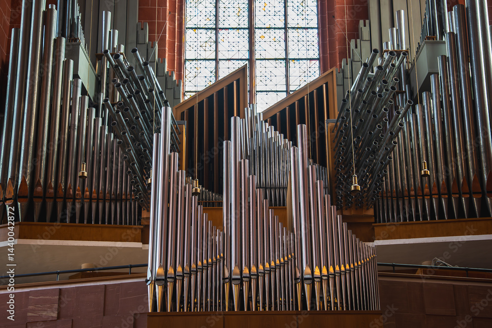 A grand pipe organ with intricate metal pipes stands before a large stained glass window. Red brick walls and wooden details highlight the historic interior.