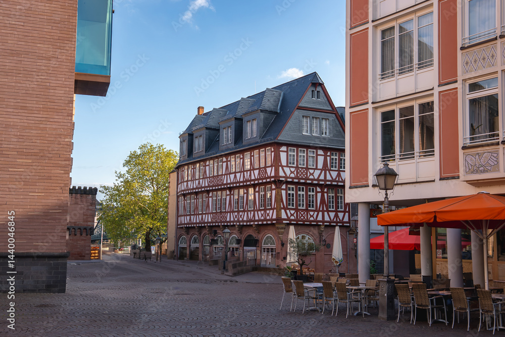 A cobblestone street in Frankfurt, Germany, with a red and white half timbered building, outdoor seating under orange umbrellas, and modern buildings nearby.