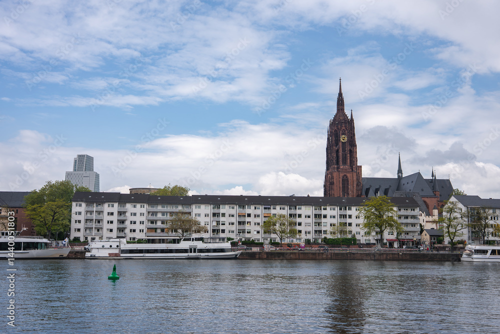 View of Frankfurt, Germany, featuring the Gothic style Frankfurt Cathedral, the Main River with moored boats, and modern high rise buildings in the background.