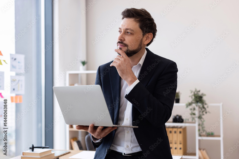 Man in early 30s wearing suit in office, holding laptop, expressing thoughtful pose, confidence. Man in early 30s wearing suit in office, holding laptop, expressing thoughtful pose, confidence.
