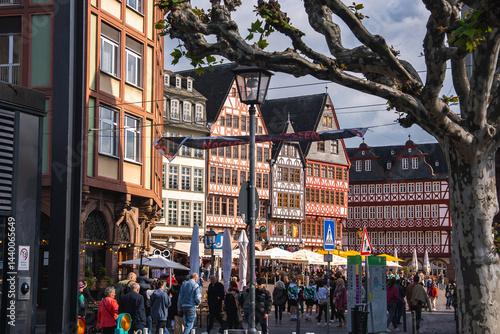 Obraz na płótnie Romerberg square in Frankfurt, Germany, features red and white half timbered buildings, a lively crowd, bare trees, street signs, and lampposts on a sunny day. w salonie