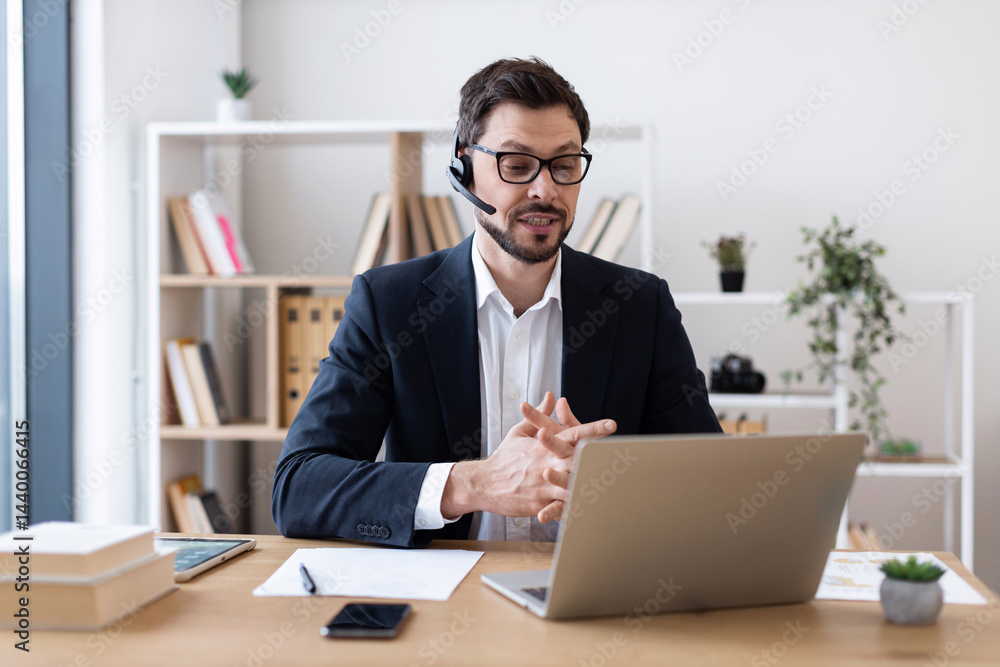 Adult man wearing suit with headset using laptop for video call in modern office. Shelves and plants in background. Adult man wearing suit with headset using laptop for video call in modern office. Shelves and plants in background.