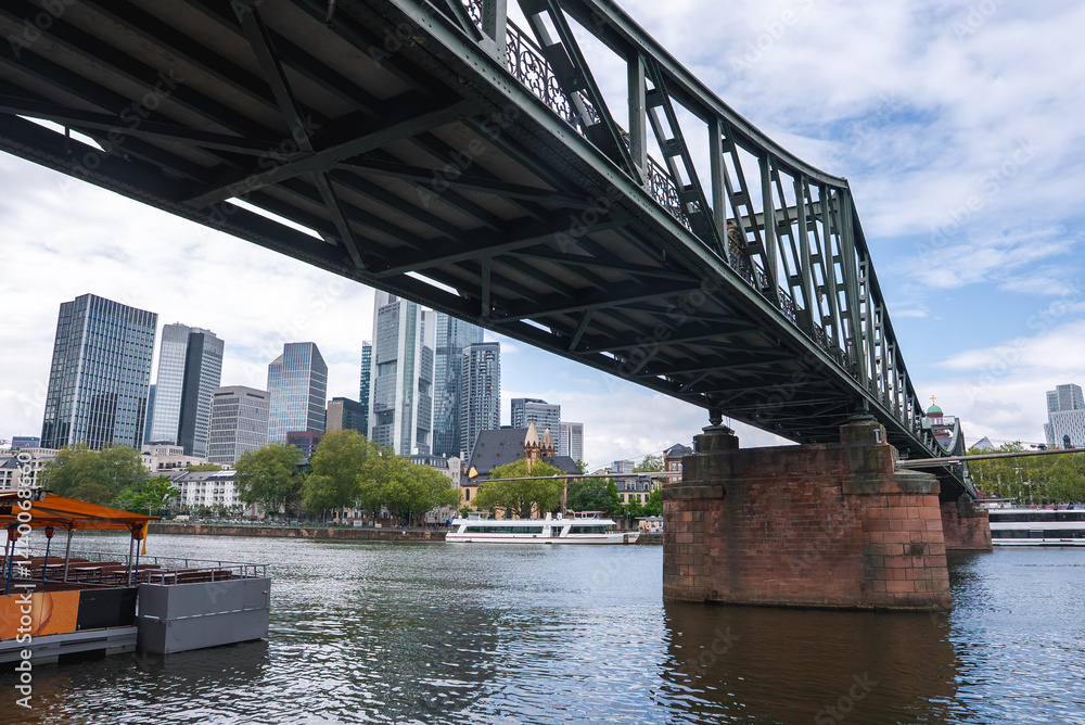The Eiserner Steg bridge spans the Main River in Frankfurt, Germany, with its green iron structure, red stone piers, and modern skyscrapers in the background.