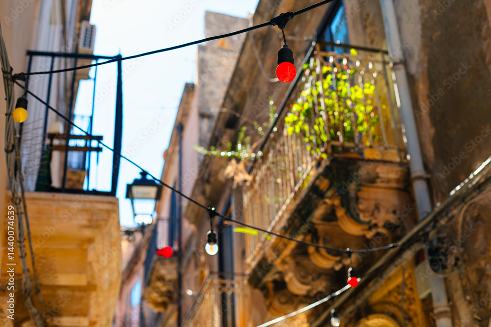 Strings of colorful lights bulbs, are strung across a narrow alley between old stone buildings with balconies. Sunlight illuminates scene, highlighting greenery on one balcony and vintage street lamp
