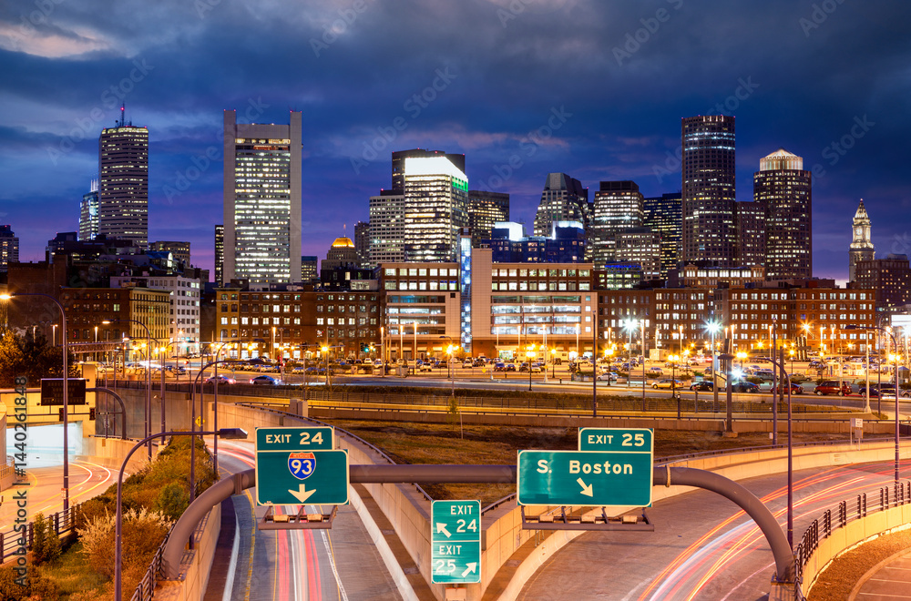 A stunning nighttime view of Boston’s illuminated skyline with bright highway lights and dramatic clouds creating a vibrant urban atmosphere.