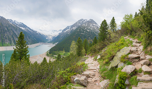 Obraz na płótnie rocky alpine trail from Schlegeis dam lake upwards. green mountain pines and grey clouds w salonie
