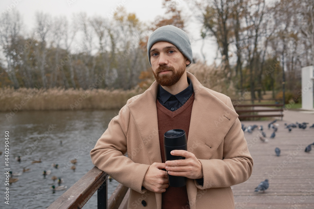 Man wearing a beige coat and hat, holding coffee by a pond in autumn.