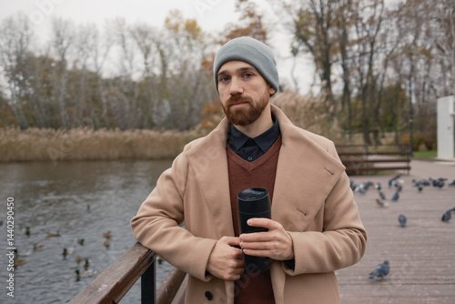 Obraz na płótnie Man wearing a beige coat and hat, holding coffee by a pond in autumn. w salonie