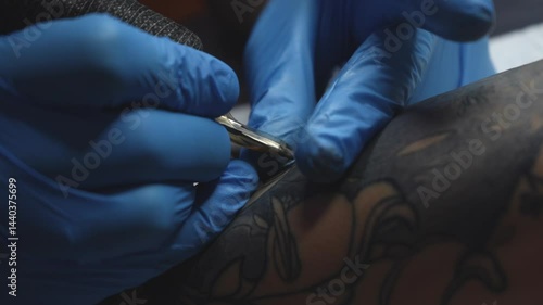 Close-up view of a tattoo artist in blue gloves creating a floral tattoo on a clients forearm. The tattoo machine is in motion as the artist outlines the design. Studio environment