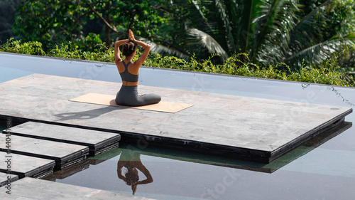 A woman practicing yoga outdoors on a mat by a serene pool, surrounded by tropical nature. She is meditating in a seated lotus position with hands raised above her head. Wellness, mindfulness.