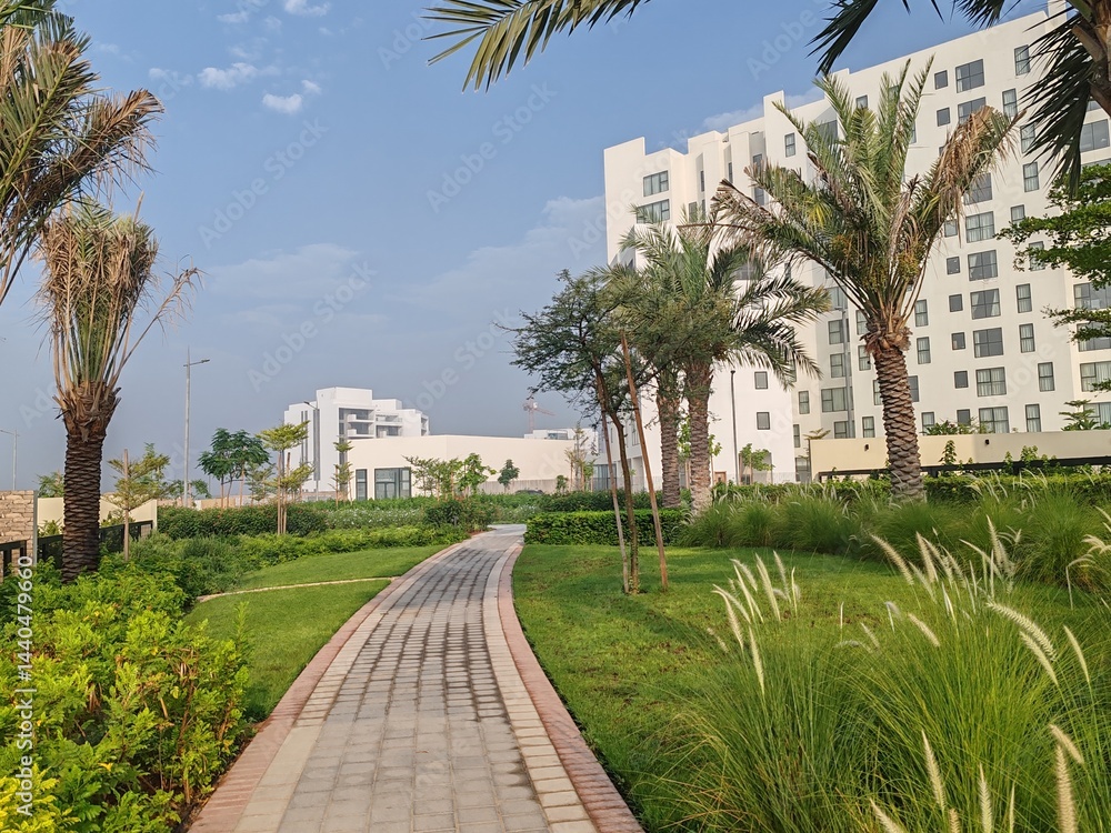 Fototapeta premium Small Interlock Path Between Landscaping Area. Green And Lush Landscaping Area With Carpet Grass And Trees Like Palm Or Washingtonia, Some Houses Or Villa's Also Beside Gardens Padestrian Pathway.