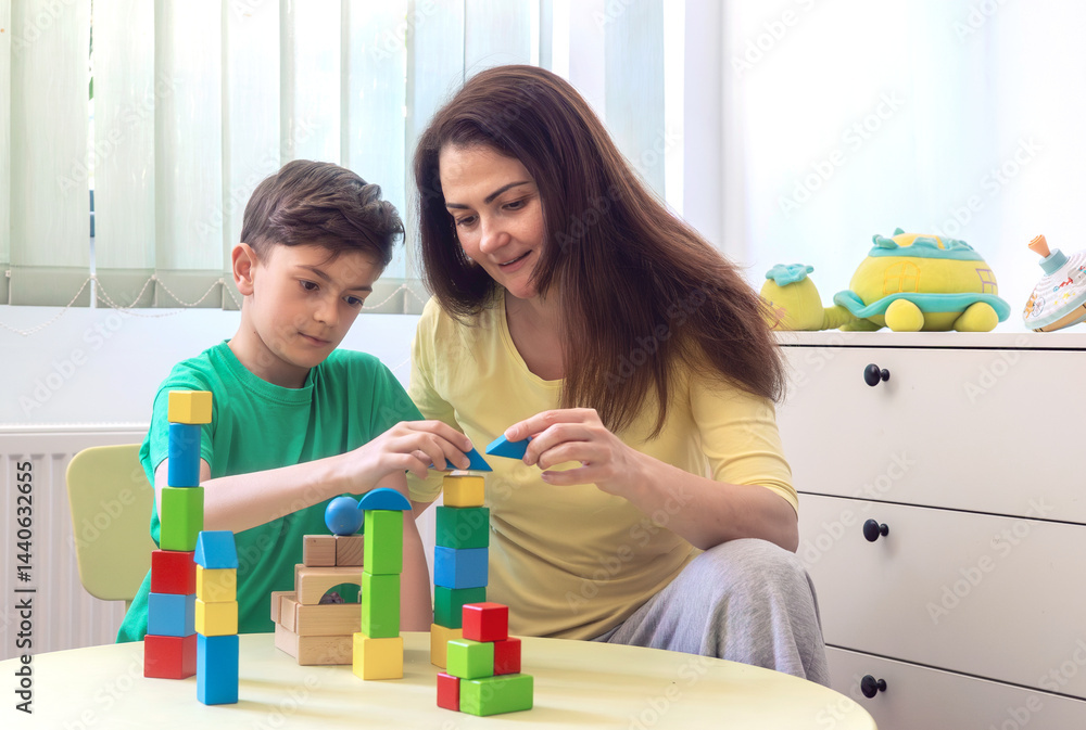 Happy young boy and woman playing with colorful wooden blocks at home. Concept of early childhood education, autism therapy, ADHD support, child psychology, occupational therapy, family connection. Happy young boy and woman playing with colorful wooden blocks at home. Concept of early childhood education, autism therapy, ADHD support, child psychology, occupational therapy, family connection.