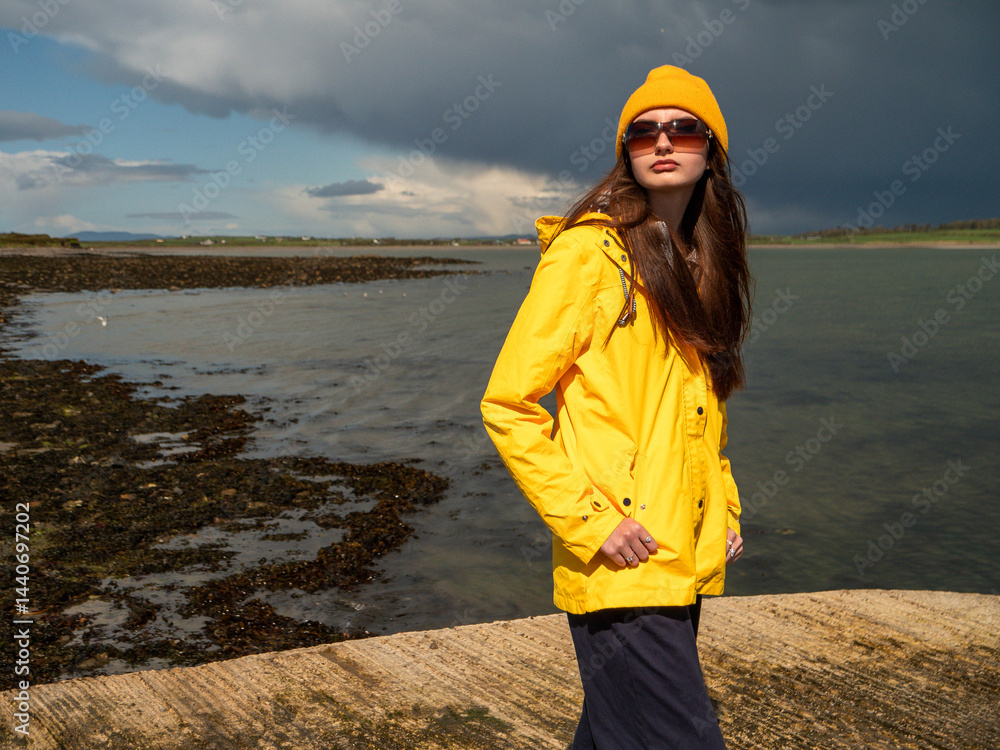 Teenager girl in yellow travel jacket standing on a pier. Huge dark storm cloud in the background. Travel in harsh weather conditions with sun and rain. County Sligo, Ireland.