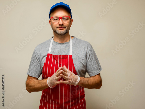 Obraz na płótnie Professional butcher in grey shirt and classic red and white stripe apron and red glasses and blue baseball cap on light color background, Hands together in plastic gloves. w salonie
