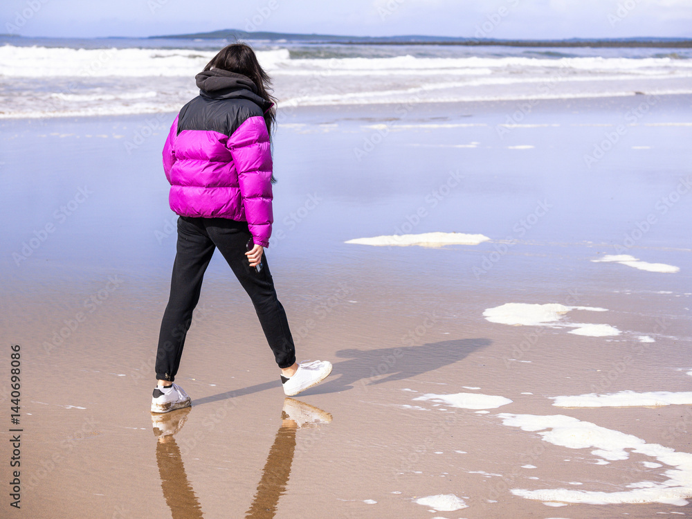Teenager girl in purple jacket walking on a wet sandy beach which reflects blue cloudy sky. Travel to seaside concept. Relaxing activity by the ocean. Selective focus.