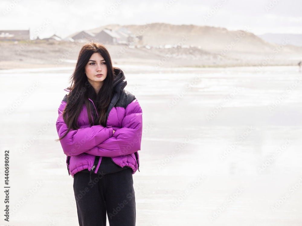 Portrait of a young teenager girl on a ocean beach with town in the background. Light and airy look. Model with long dark hair in purple jacket hands crossed, and sad face expression.