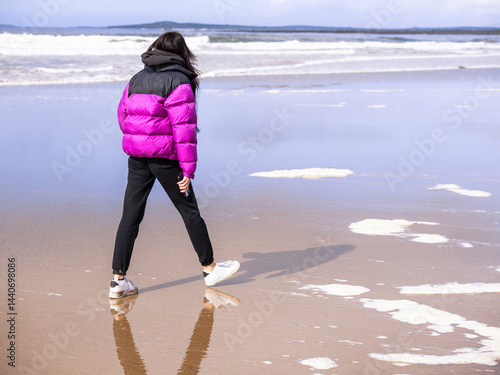 Obraz na płótnie Teenager girl in purple jacket walking on a wet sandy beach which reflects blue cloudy sky. Travel to seaside concept. Relaxing activity by the ocean. Selective focus. w salonie