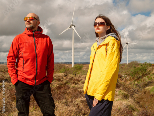 Obraz na płótnie Father in red jacket and daughter in yellow enjoy warm sunny day. Wind turbine and huge wild field and cloudy sky in the background. Divorced man day with his daughter concept. Outdoor activity. w salonie