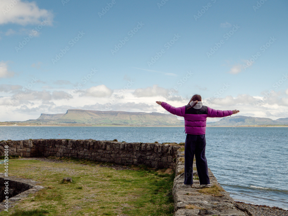 Obraz premium Girl tourist standing on a pier with her hands up in the air. Famous Benbulbin flat top mountain and ocean in the background. Travel in Ireland. Enjoy amazing Irish nature scenery concept.
