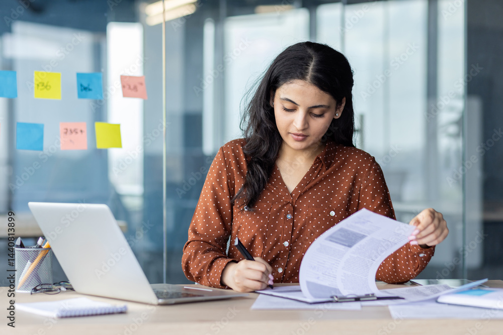 A focused woman, working intently on paperwork at a desk in a modern office setting. A laptop sits beside her, and sticky notes are visible.
