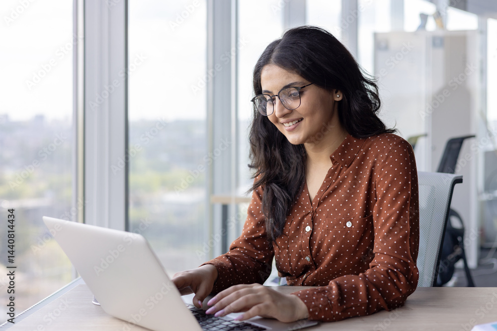 A smiling woman with glasses works on a laptop in a modern office setting. The office has a large window with a city view in the background.