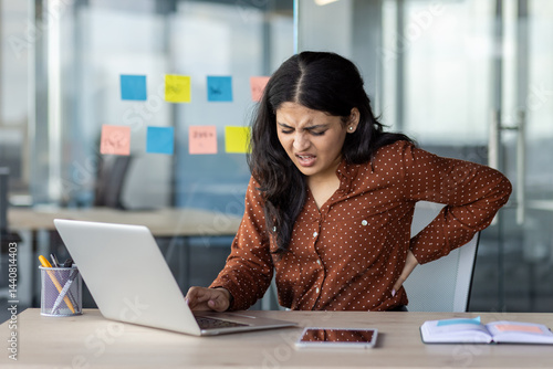 An indian woman in pain clutching her lower back while sitting at a desk in an office with a laptop open in front of her.