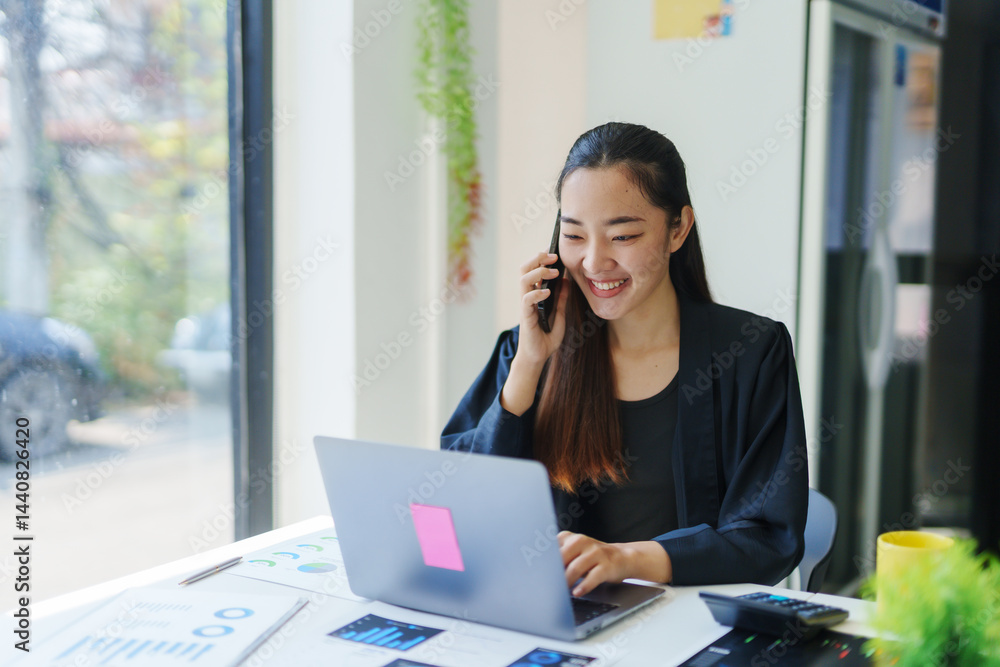 Obraz premium Smiling businesswoman working on a laptop while talking on a smartphone in a bright, modern office space filled with natural light