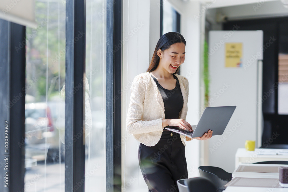 Smiling Asian businesswoman using a laptop while standing beside a window in a modern office, enjoying a productive workday Smiling Asian businesswoman using a laptop while standing beside a window in a modern office, enjoying a productive workday