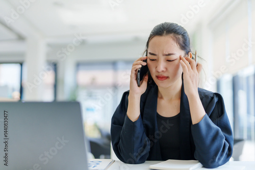 Asian manager feeling tired and frustrated after phone call with client, touching temples to relieve headache, sitting at desk with laptop and documents