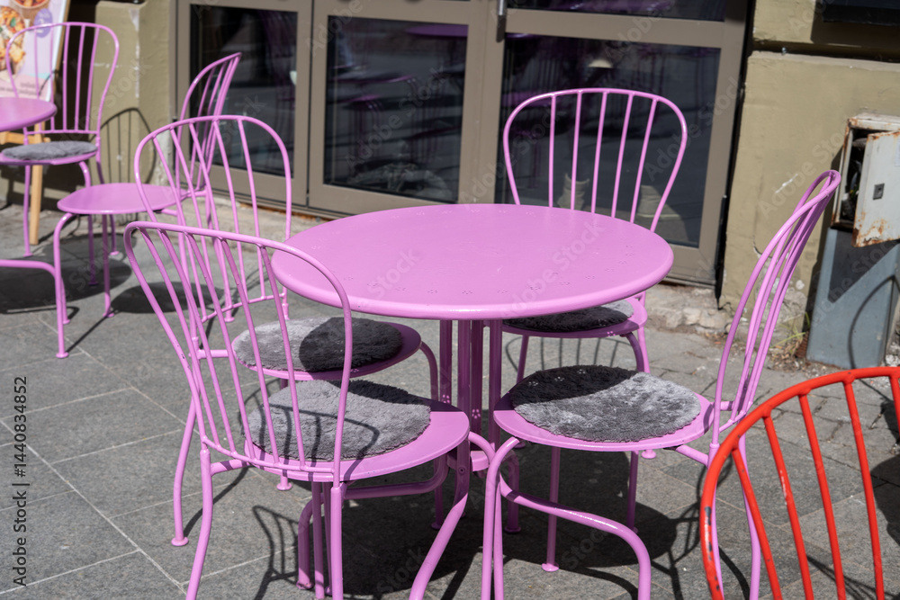 sunlit pastel sidewalk caf scene featuring a round pink metal table surrounded by matching lavender chairs with gray cushions, set against an urban storefront, evoking relaxed outdoor leisure and vibr