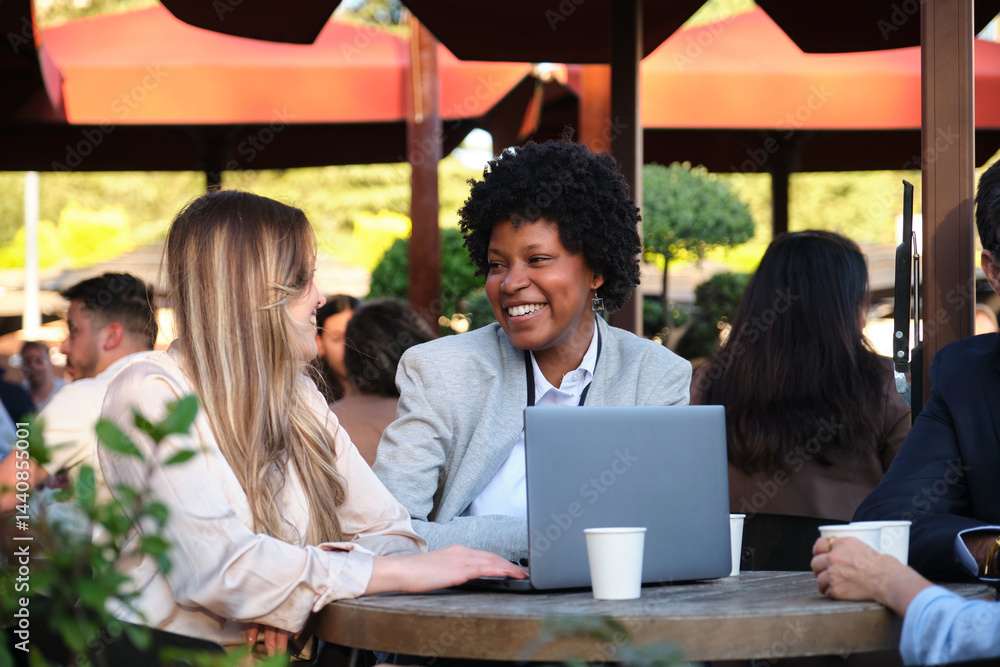 Two businesswomen having a coffee break, working on a laptop and discussing at a table of an outdoor cafe