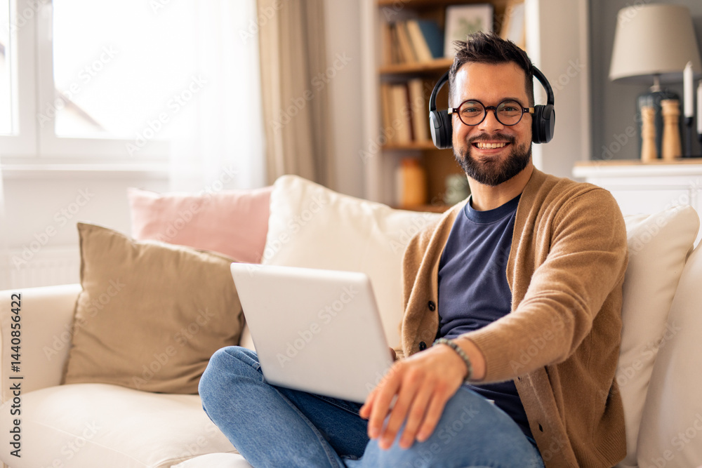 Smiling Caucasian Man Wearing Headphones Working on Laptop in a Bright Cozy Living Room Smiling Caucasian Man Wearing Headphones Working on Laptop in a Bright Cozy Living Room
