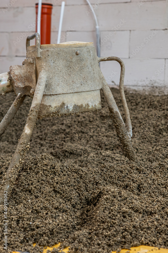 A construction site featuring a concrete mixer on gravel, a white brick wall with visible pipes, including an orange pipe, in the background.