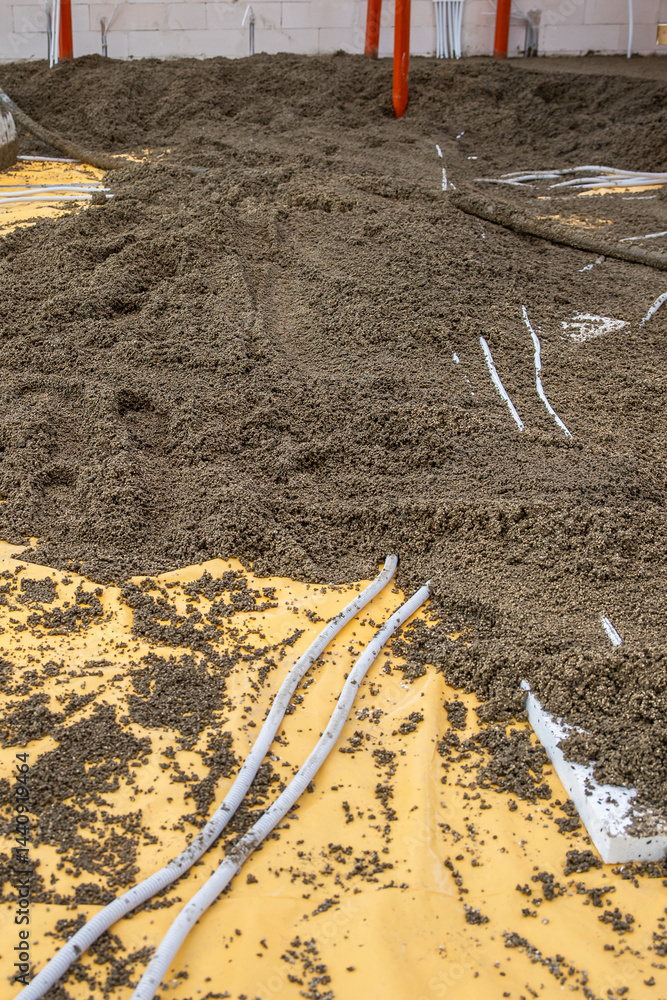 Indoor construction site floor in progress with yellow vapor barrier, loose aggregate, white conduits, orange supports, and a white block wall.