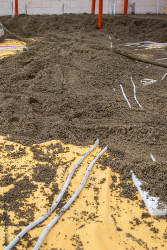 Indoor construction site floor in progress with yellow vapor barrier, loose aggregate, white conduits, orange supports, and a white block wall.