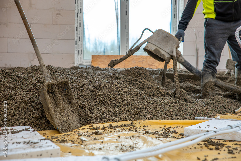 Construction workers pour and level concrete over yellow plastic sheeting and insulation panels, with tools and bare trees visible through window openings.