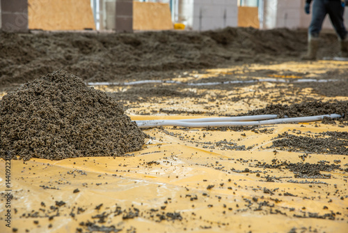 A construction site featuring a gravel pile on a yellow plastic sheet, pipes arranged for foundation work, and partially built concrete walls.