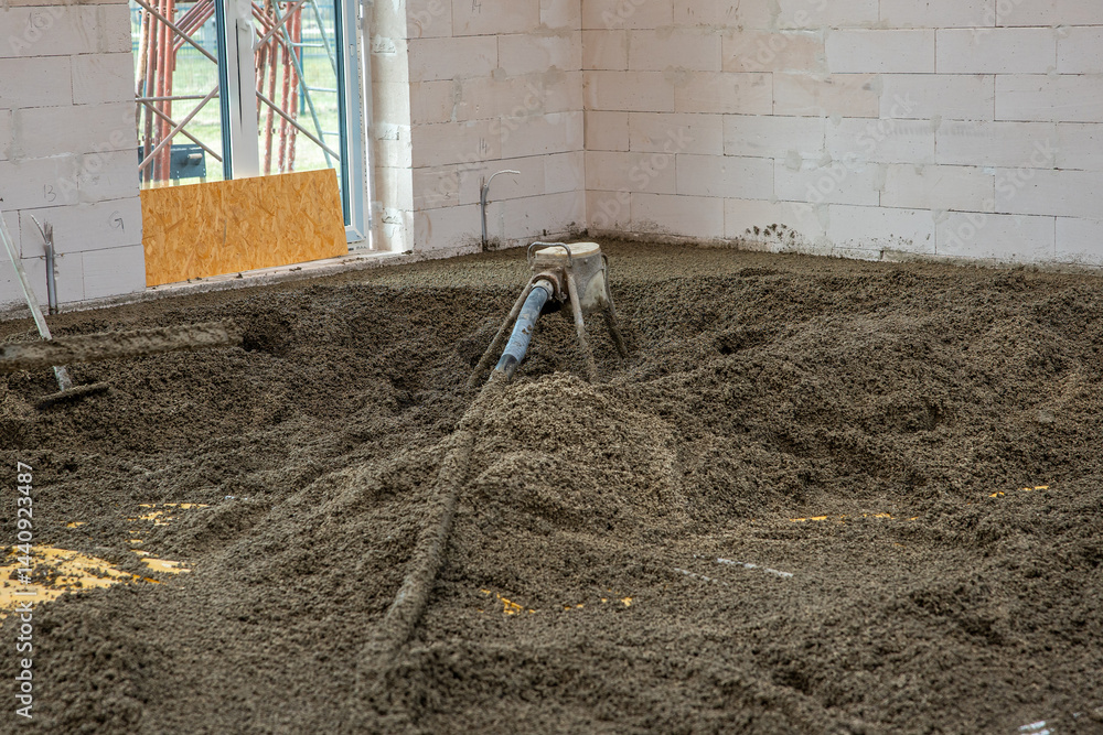 White brick walls and an unfinished floor being leveled with concrete. A hose distributes material, with light entering through a window opening.