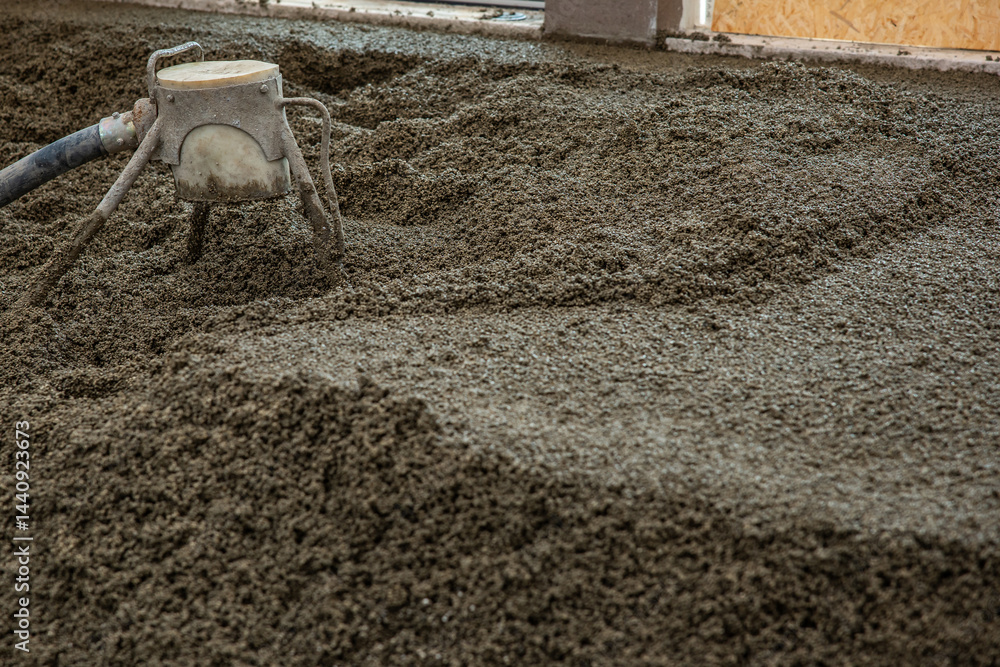 Freshly poured concrete floor in an indoor construction site, with a leveling tool connected to a hose in use. Partially visible wall and window frame.