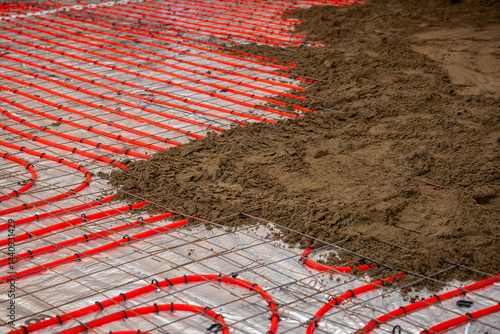 Red underfloor heating pipes are installed over reflective insulation, secured by a metal grid, with Estrich concrete partially covering the pipes.