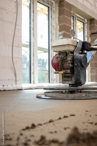 Interior of a construction site showing a power trowel smoothing concrete flooring over underfloor heating pipes, with brick walls and large windows.