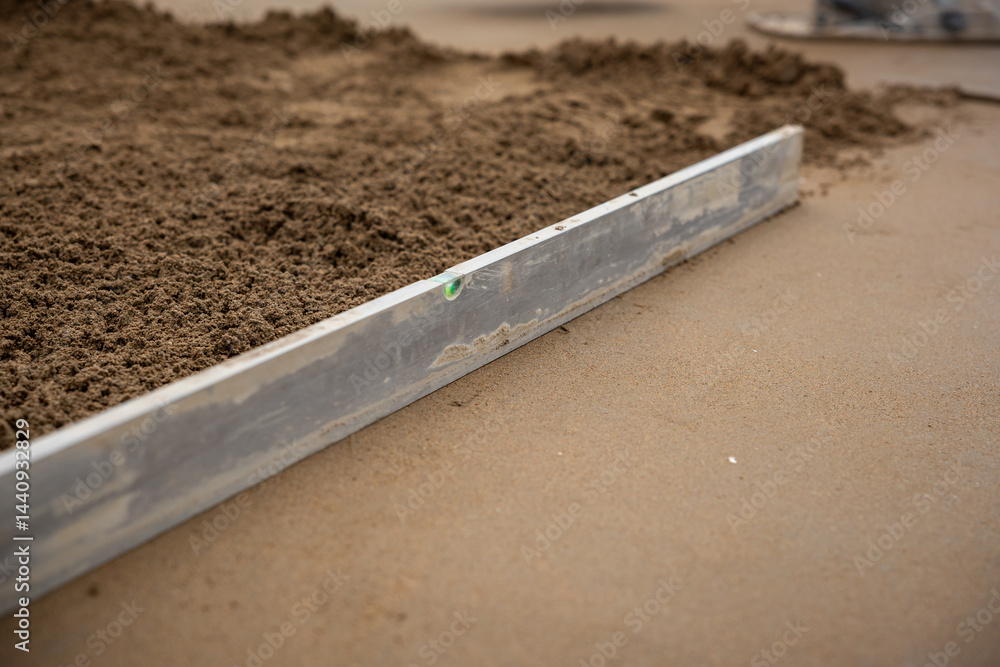 A metal screed tool with a green bubble level levels wet concrete, showing smooth and rough textures during a flooring installation process.